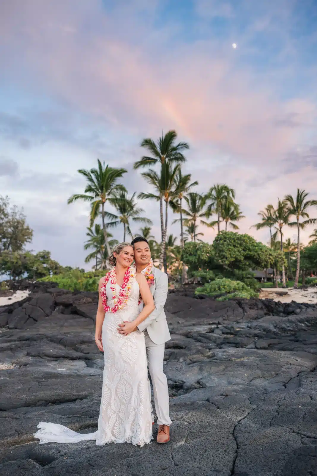 Couple on the lava with a rainbow and the moon above them after their Big Island wedding